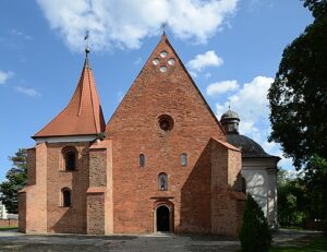 Ceglany kościół z charakterystyczną, dwuspadową fasadą i strzelistą wieżą na tle błękitnego nieba. Po prawej stronie widoczna jest przyległa kaplica o jasnej elewacji i kopułowym dachu.