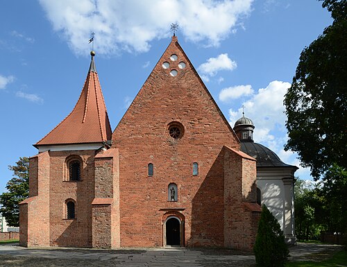 Ceglany kościół z charakterystyczną, dwuspadową fasadą i strzelistą wieżą na tle błękitnego nieba. Po prawej stronie widoczna jest przyległa kaplica o jasnej elewacji i kopułowym dachu.