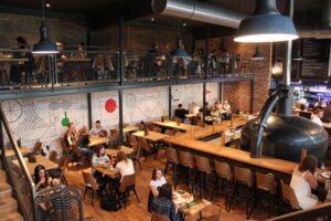 Wnętrze nowoczesnej restauracji z antresolą, gdzie goście siedzą przy drewnianych stolikach, a ściany zdobią industrialne dekoracje. Na pierwszym planie widoczny jest duży zbiornik, a w tle znajduje się bar z menu na tablicy.