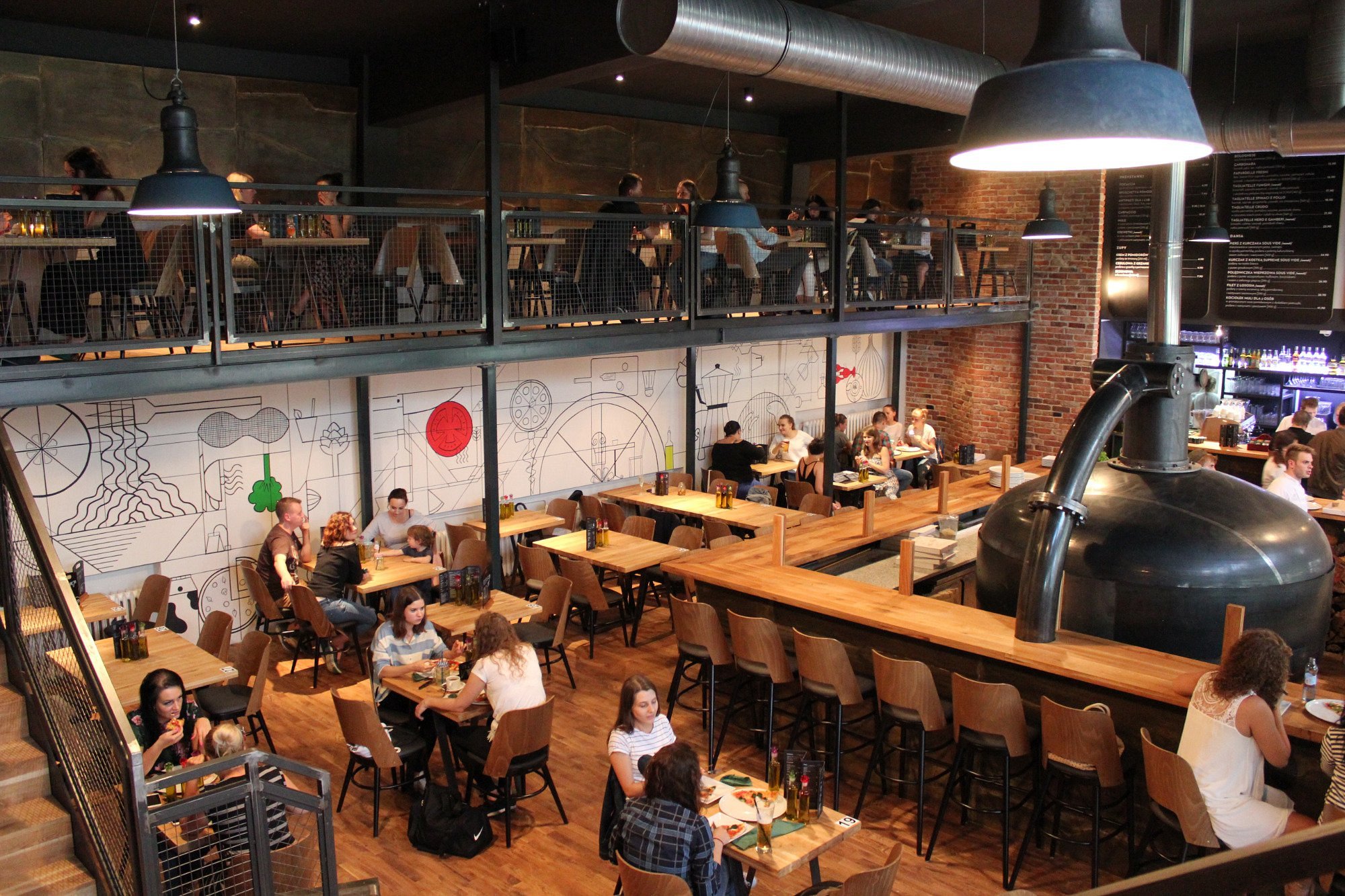 Wnętrze nowoczesnej restauracji z antresolą, gdzie goście siedzą przy drewnianych stolikach, a ściany zdobią industrialne dekoracje. Na pierwszym planie widoczny jest duży zbiornik, a w tle znajduje się bar z menu na tablicy.