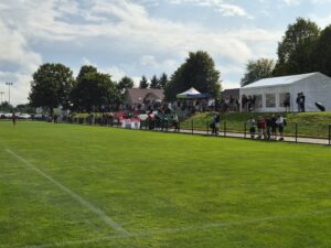 Na boisku sportowym z trawiastą nawierzchnią zgromadziło się wielu widzów, którzy stoją obok białych namiotów i trybun. W tle znajdują się drzewa oraz kilka budynków, co sugeruje otoczenie parku lub miejskiej przestrzeni rekreacyjnej.