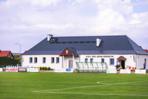 Budynek klubu sportowego z napisem "K.K.S. 1922 Lechia Kostrzyn" na tle boiska piłkarskiego otoczonego ławkami i reklamami. Na boisku widoczna jest osłonięta ławka dla zawodników rezerwowych, a w tle stoi dom o nowoczesnym designie.
